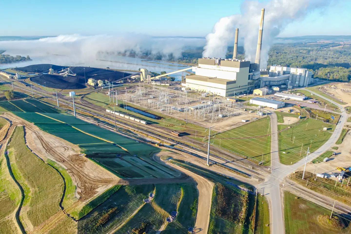 Aerial view of a power plant beside a river on a sunny morning: a large black coal pile and a coal train on the left, a pale boiler building with two tall stacks giving off white plumes on the right, and green fields and roads in the foreground.