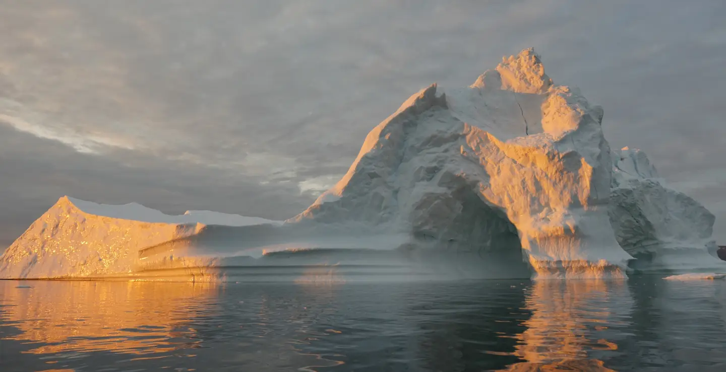A large iceberg with steep, jagged peaks floating on calm, dark water under a cloudy sky, its left side lit golden by a low sun and reflected in the water.