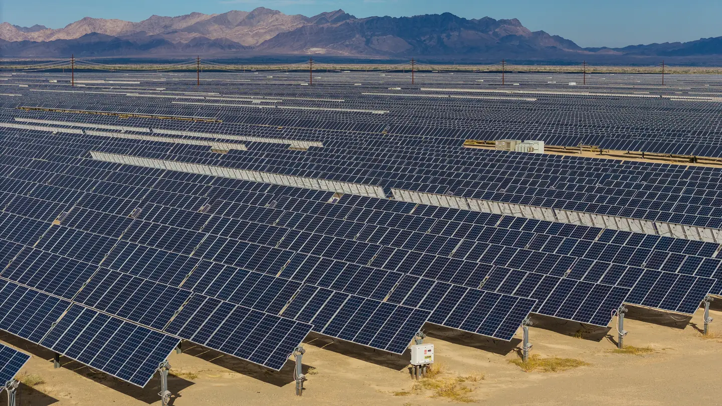 Long rows of tilted solar panels covering flat desert ground, with a line of power poles and bare mountains in the background under a clear blue sky.